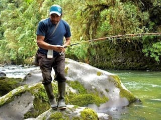 Eladio standing on mossy river rocks while fishing, surrounded by lush forest and clear green water.