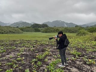 Jhonny photographing birds in a misty field with a telephoto lens, mountains in the background, wearing a black jacket and backpack.