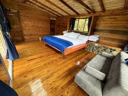 Spacious wooden cabin room with king bed, large window, and sofa at Cabañas Tamiaju