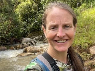 Toni smiling by a rushing creek in the forest, wearing camouflage and a backpack.