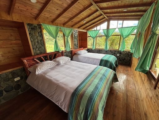 Wide-angle view of a rustic cabin interior at Cabañas Tamiaju with two beds, vibrant striped blankets, wood floors, and wraparound windows.