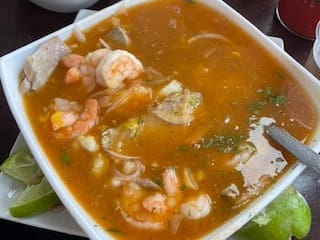 A steaming bowl of Ecuadorian encebollado, a hearty fish stew with shrimp, yuca, onions, and lime wedges on the side.